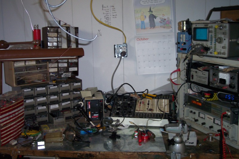 workspace.  To the left is a server rack with electronics on it.  To the right is a collection of small trays.  In the center is a soldering station and a large prototyping box